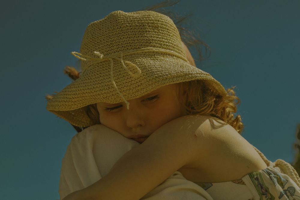 Figure in a straw sunhat rests against their knees in warm summer lighting against a blue sky.