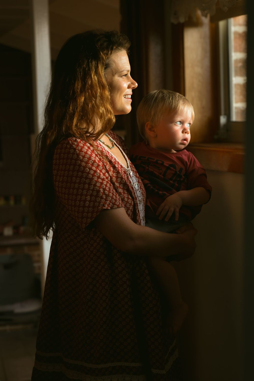 A parent and child in matching plaid shirts stand by a window in warm, natural lighting.