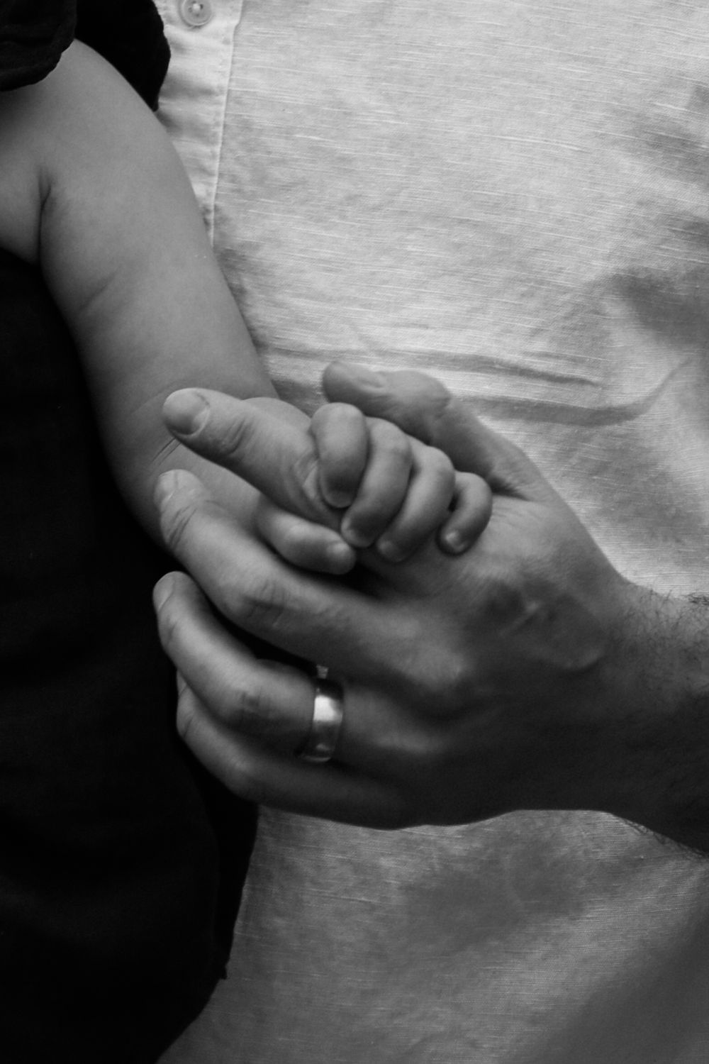 Black and white close-up photo of a baby's tiny hand holding an adult's finger with a wedding ring.