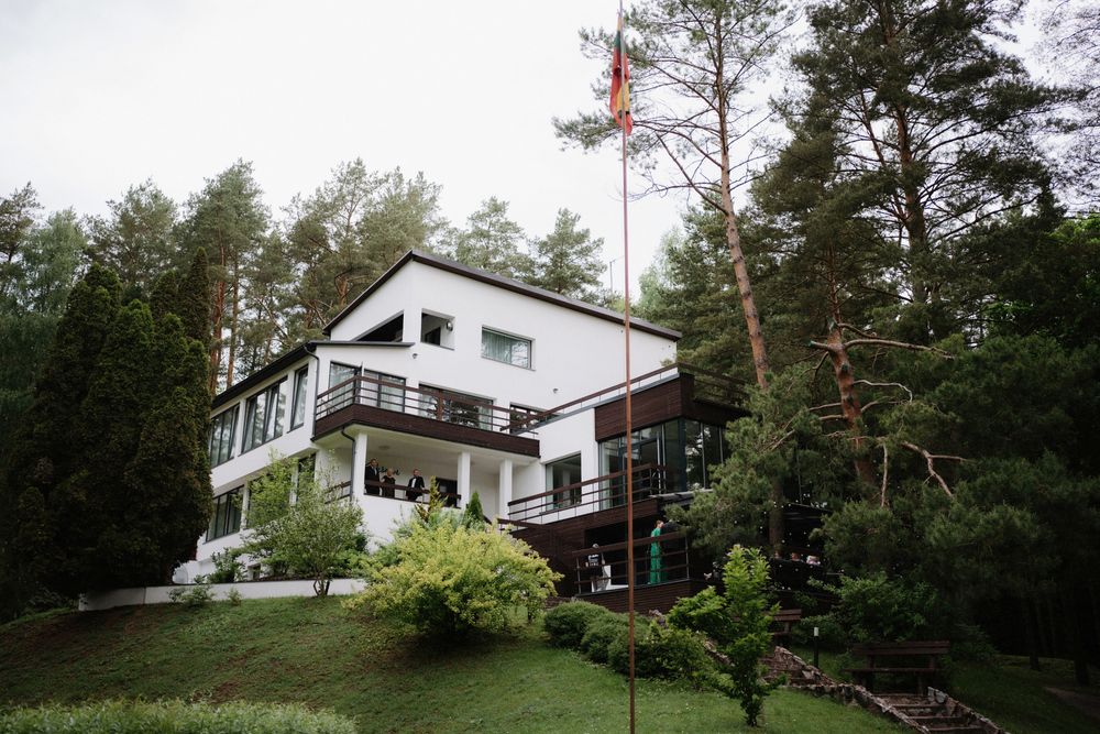 Modern white house with large windows nestled among pine trees on a hillside.