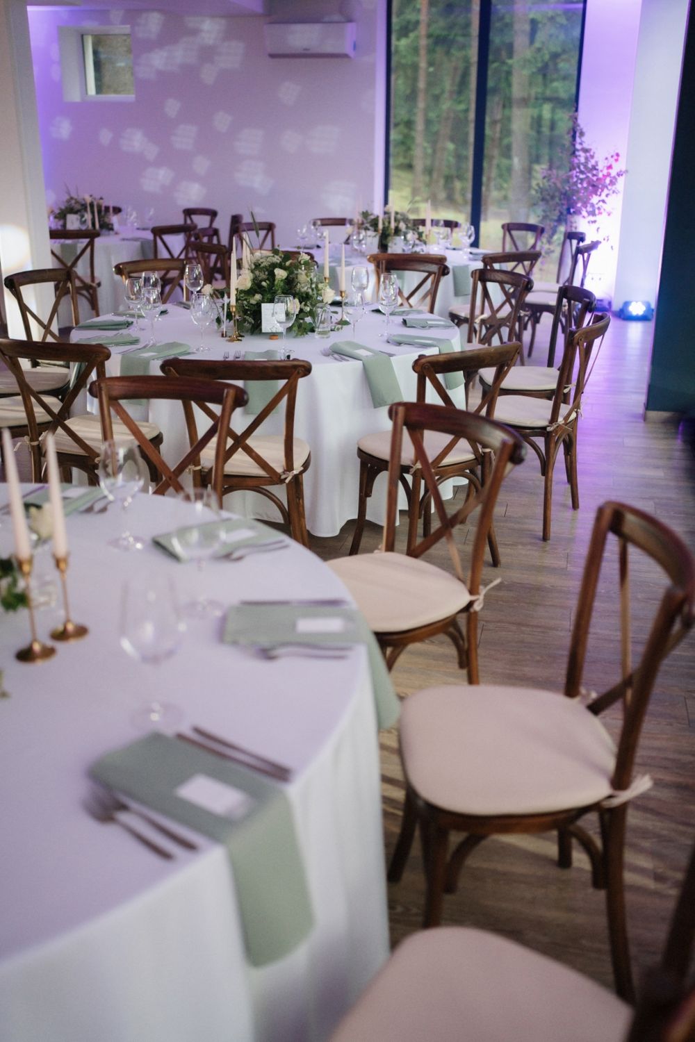 Elegant wedding reception venue with rustic wooden chairs and white tablecloths illuminated by purple ambient lighting.