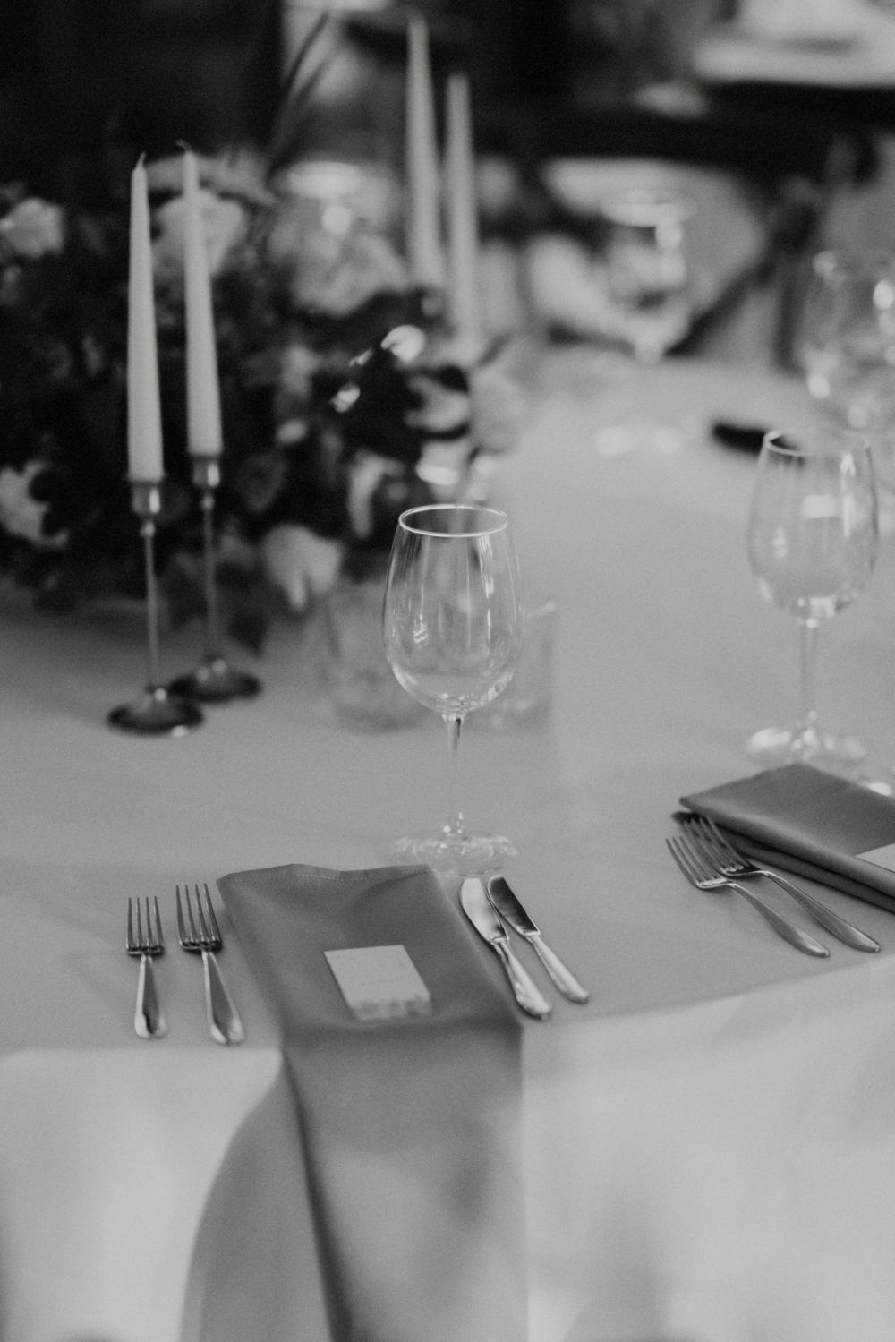 Black and white close-up of formal table setting with wine glasses, candles and silverware.