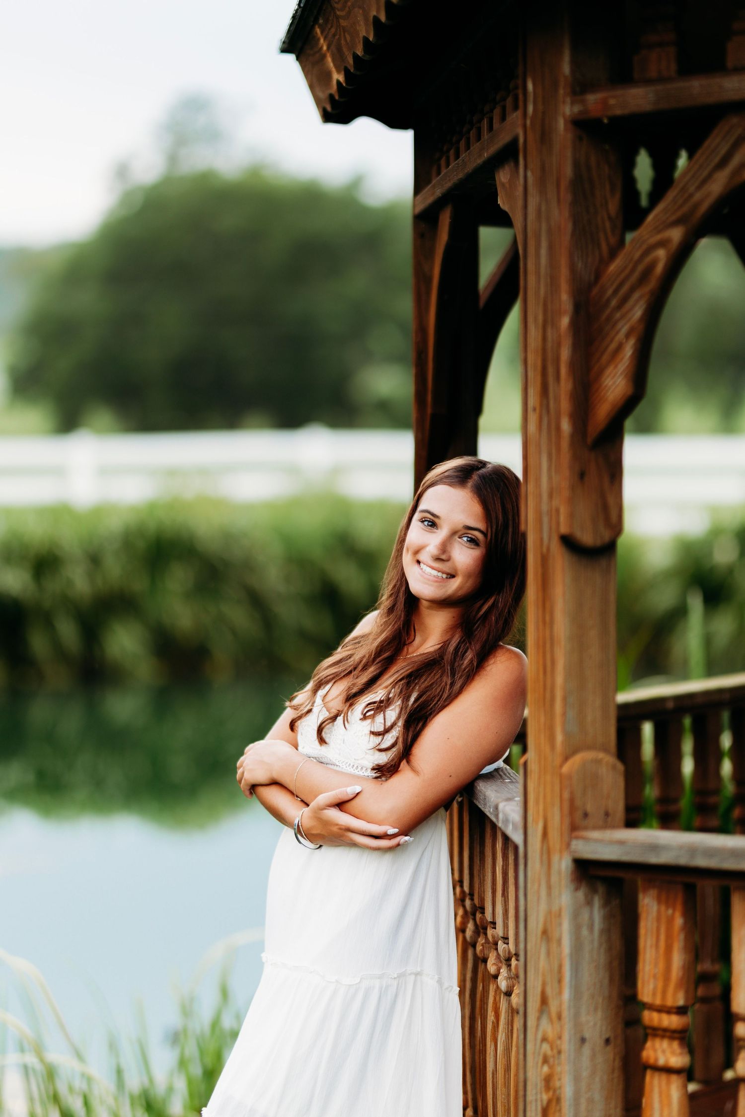 Young woman in white dress leaning against wooden gazebo post near a peaceful pond.