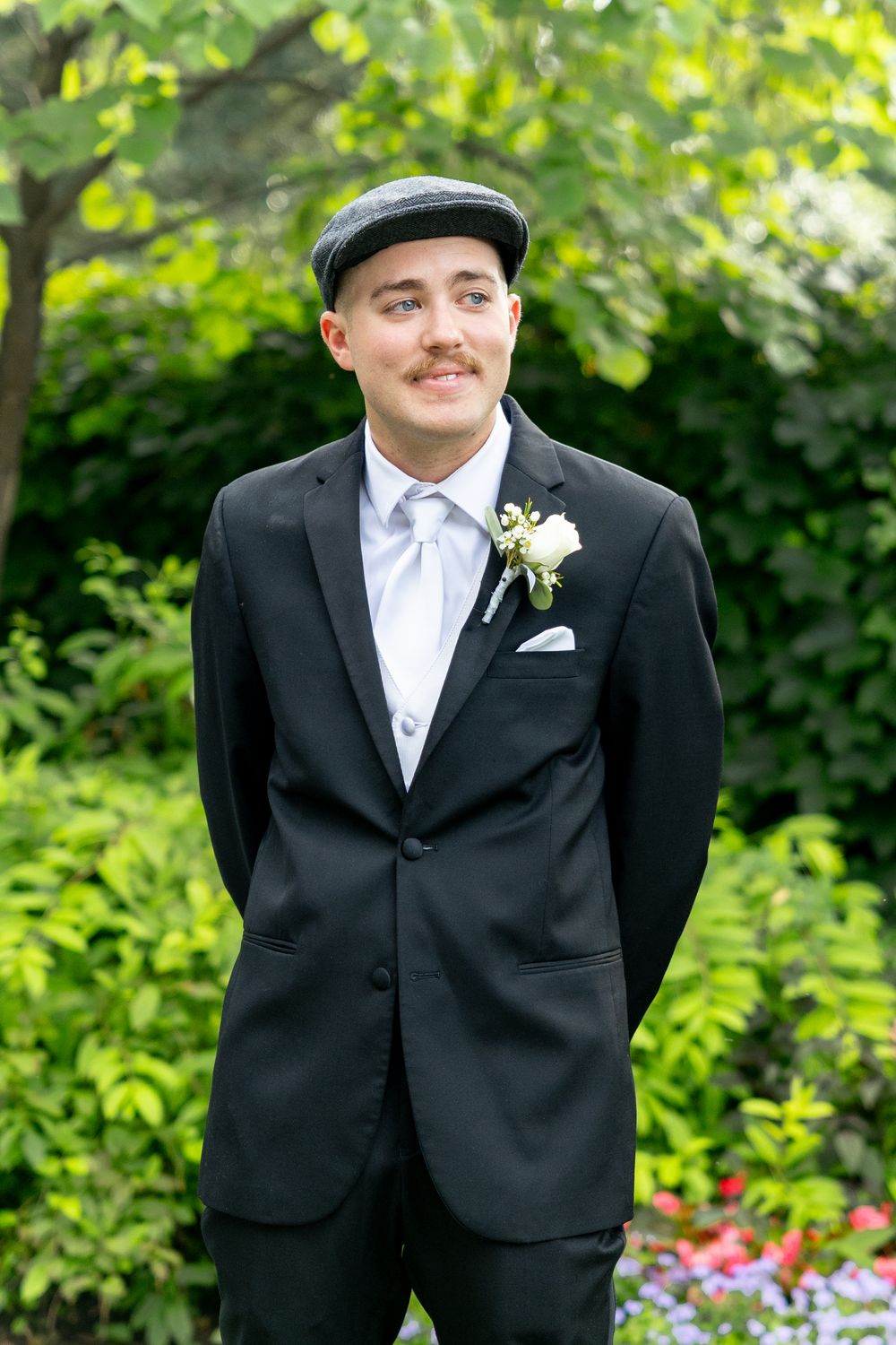 A person in a formal black tuxedo and white tie stands outdoors against a green garden background.