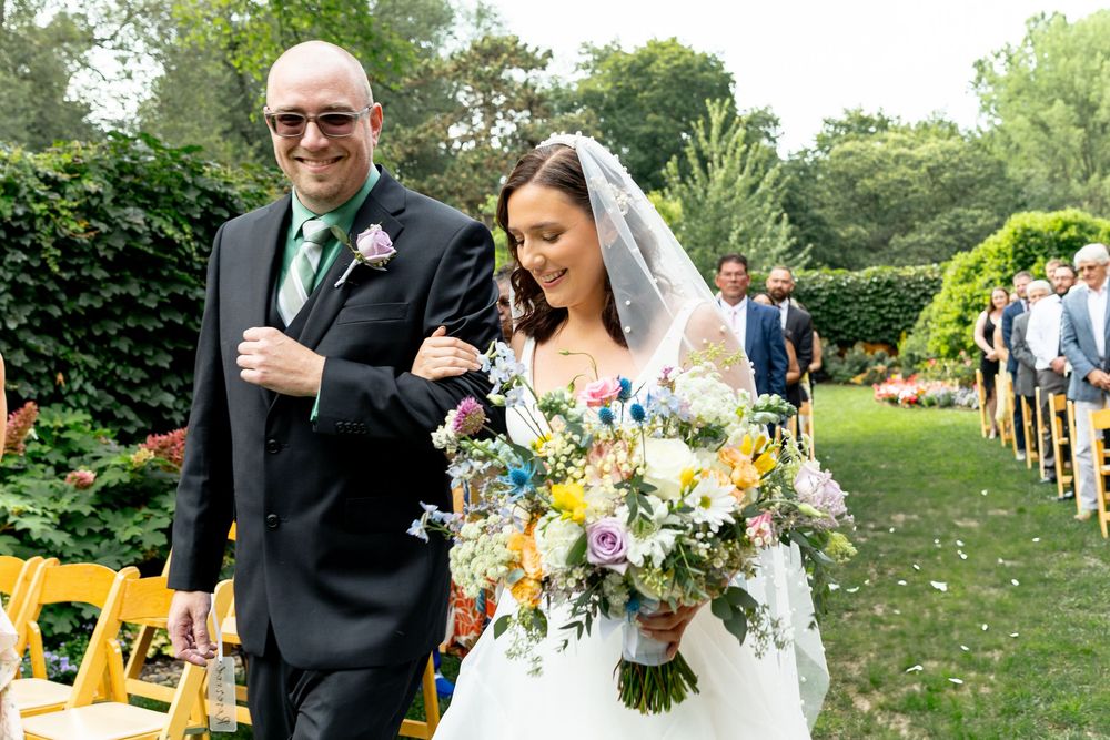 A wedding ceremony processional in a lush garden setting with yellow chairs and floral bouquets.