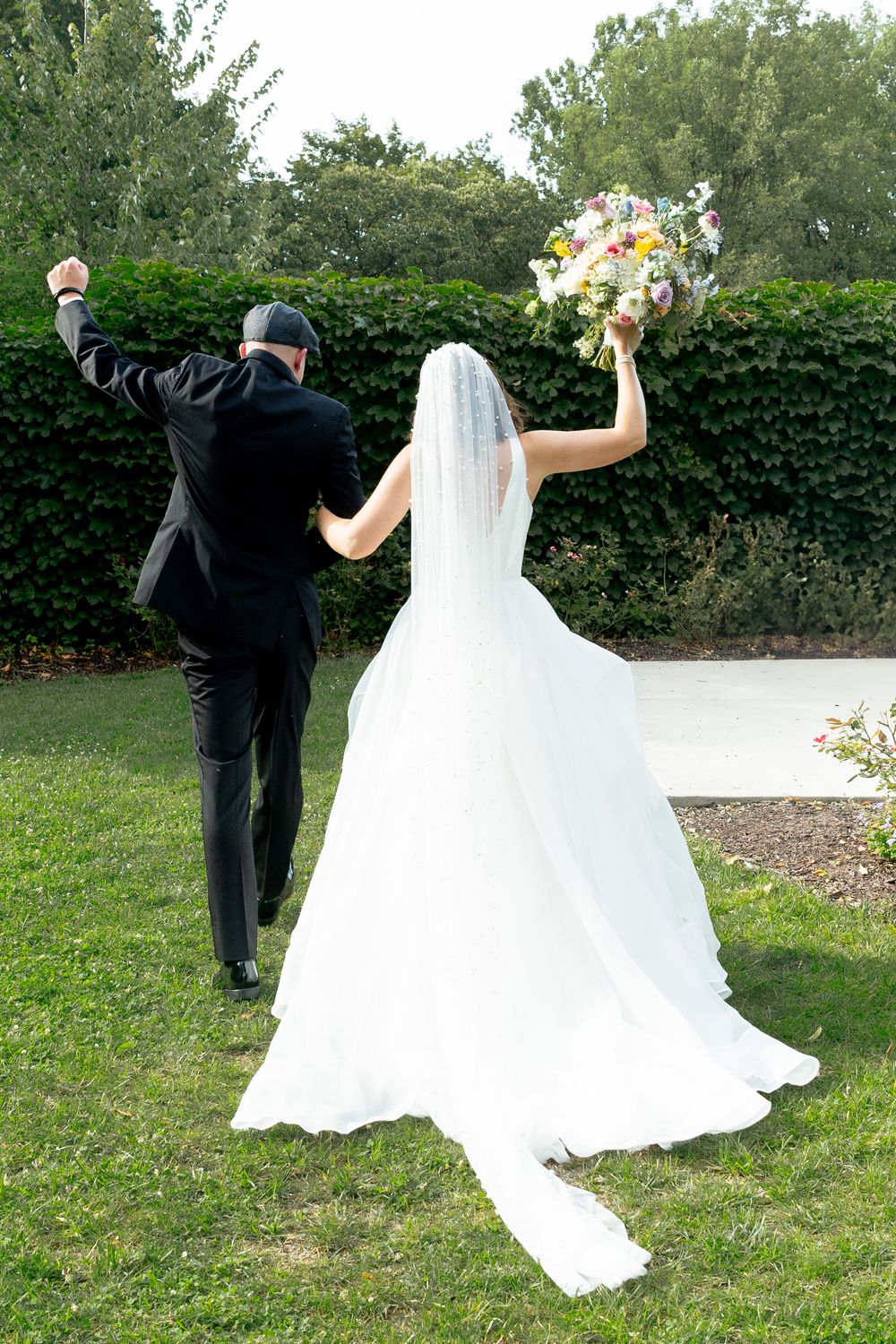 A celebratory exit shot of newlyweds raising their arms in joy against a hedge backdrop.