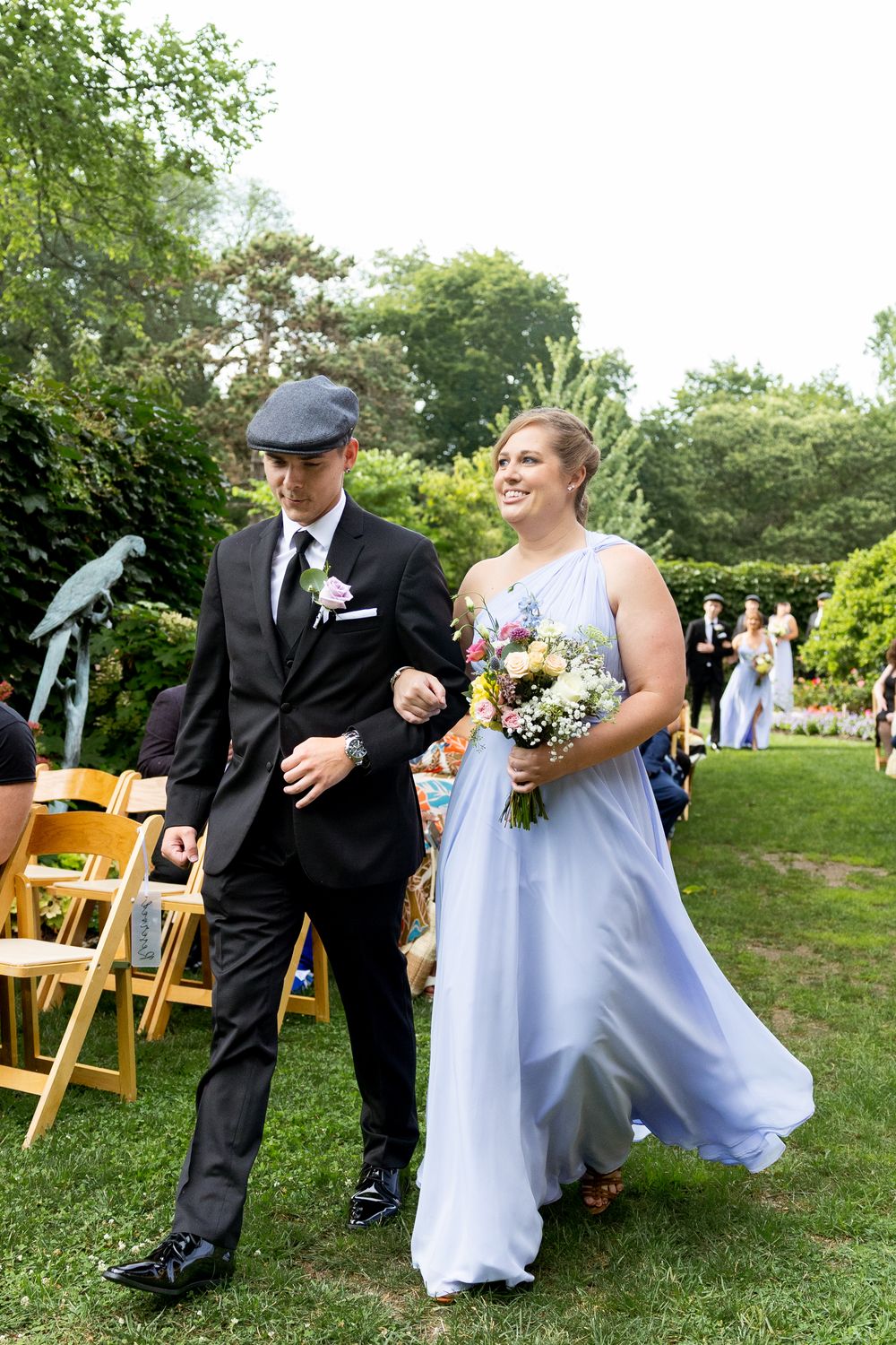 A couple walks down the aisle at an outdoor garden wedding ceremony with yellow chairs and greenery.