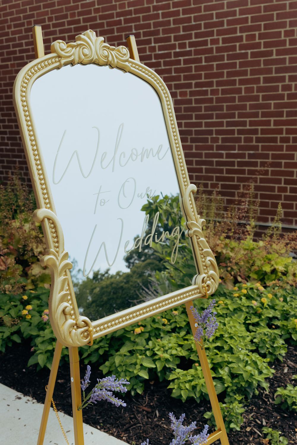 An ornate gold-framed mirror with 'Welcome' text stands on an easel against a brick wall surrounded by green shrubbery.