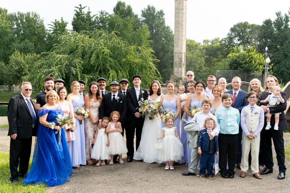 Large wedding party group photo with bridesmaids in blue dresses and flower girls in white gathered outdoors.
