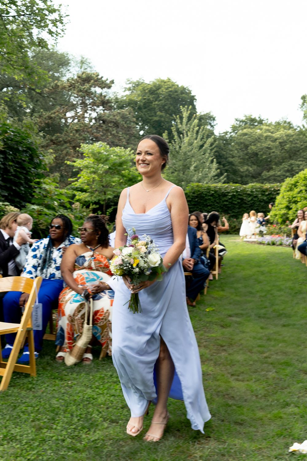 A bridesmaid in a flowing light blue dress walks down an outdoor garden aisle carrying a bouquet at a wedding ceremony.