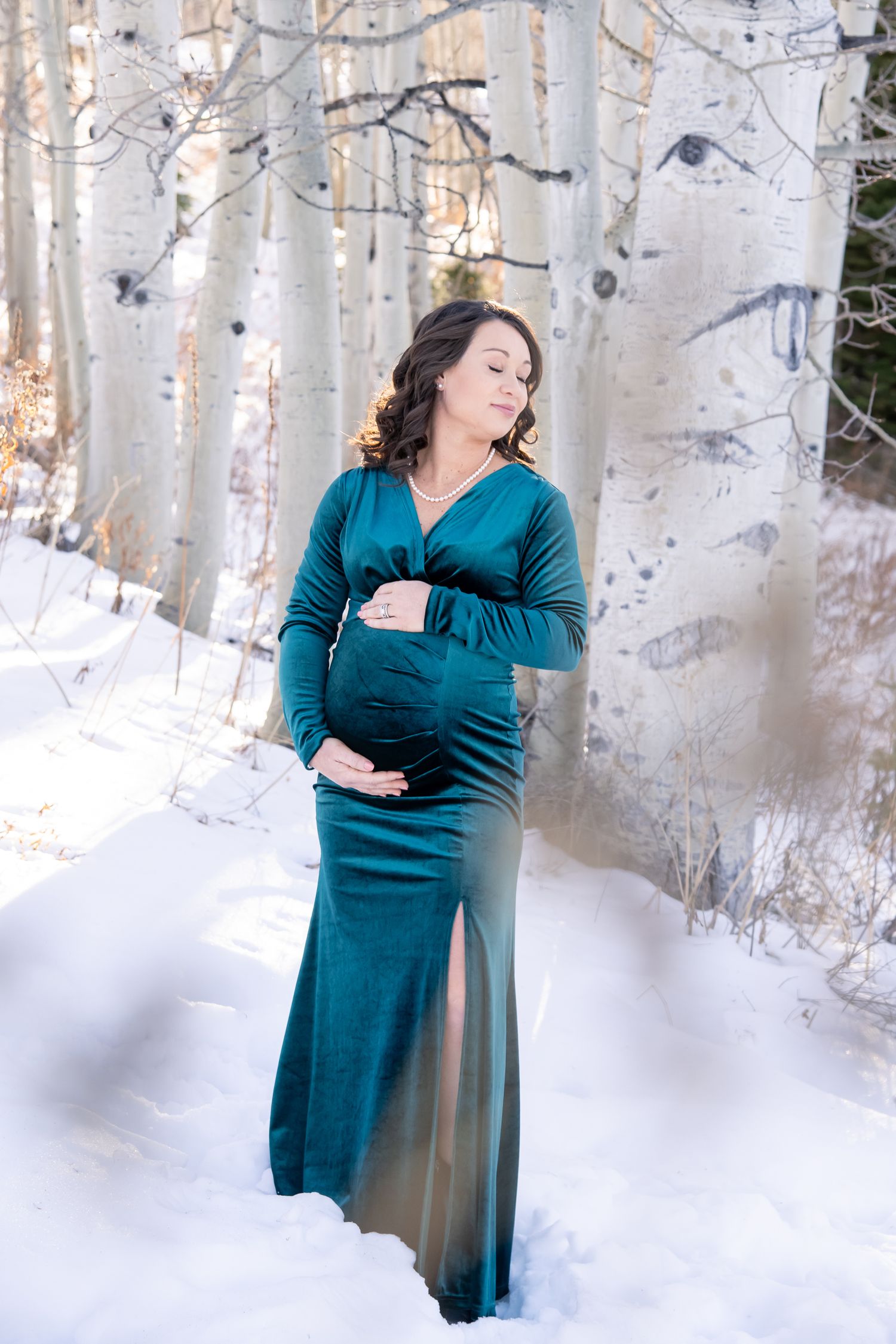 A woman in a teal wrap dress poses elegantly among snow-covered aspen trees in a winter forest setting during maternity photos in Aspen CO.