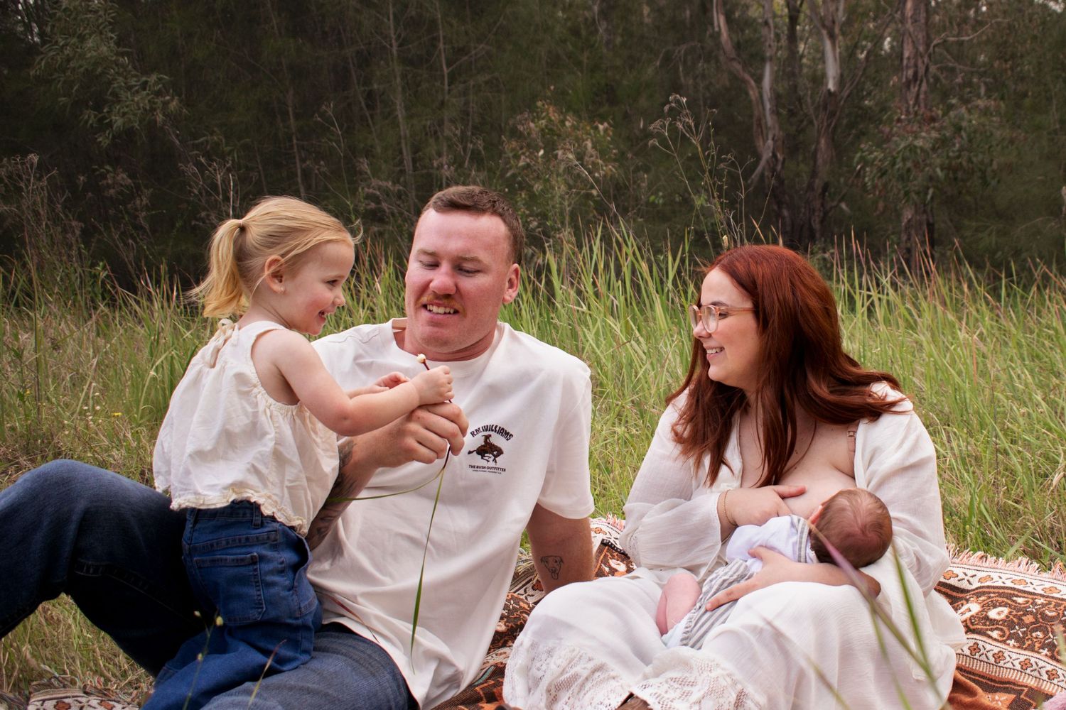 toddler playing with dad and mum as mum breastfeeds newborn, gathered on a picnic rug in a grassy field in Maitland NSW newborn photoshoot