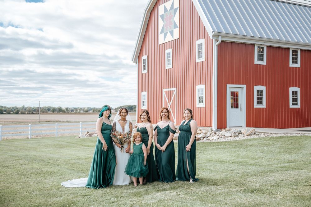 A bridal party in emerald green dresses poses in front of a classic red barn.