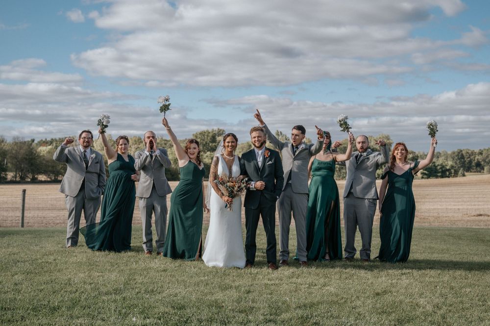 A wedding party in green and gray attire celebrates on a field with a barn in the background.