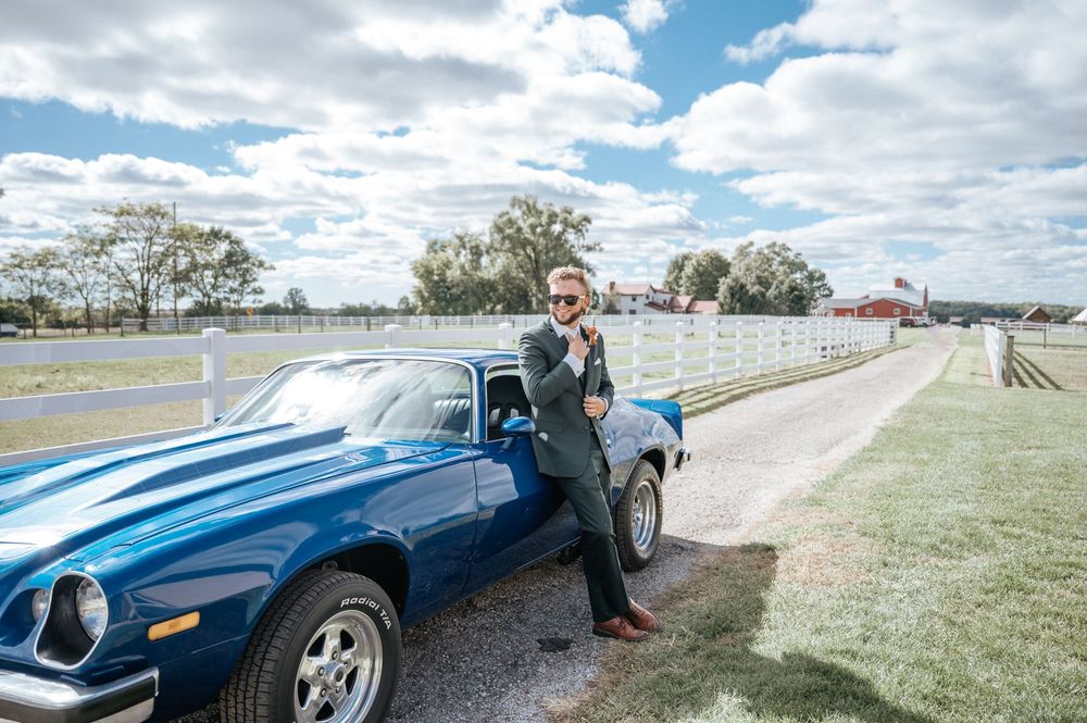 A groom in a suit poses next to a classic blue vintage car on a sunny day.