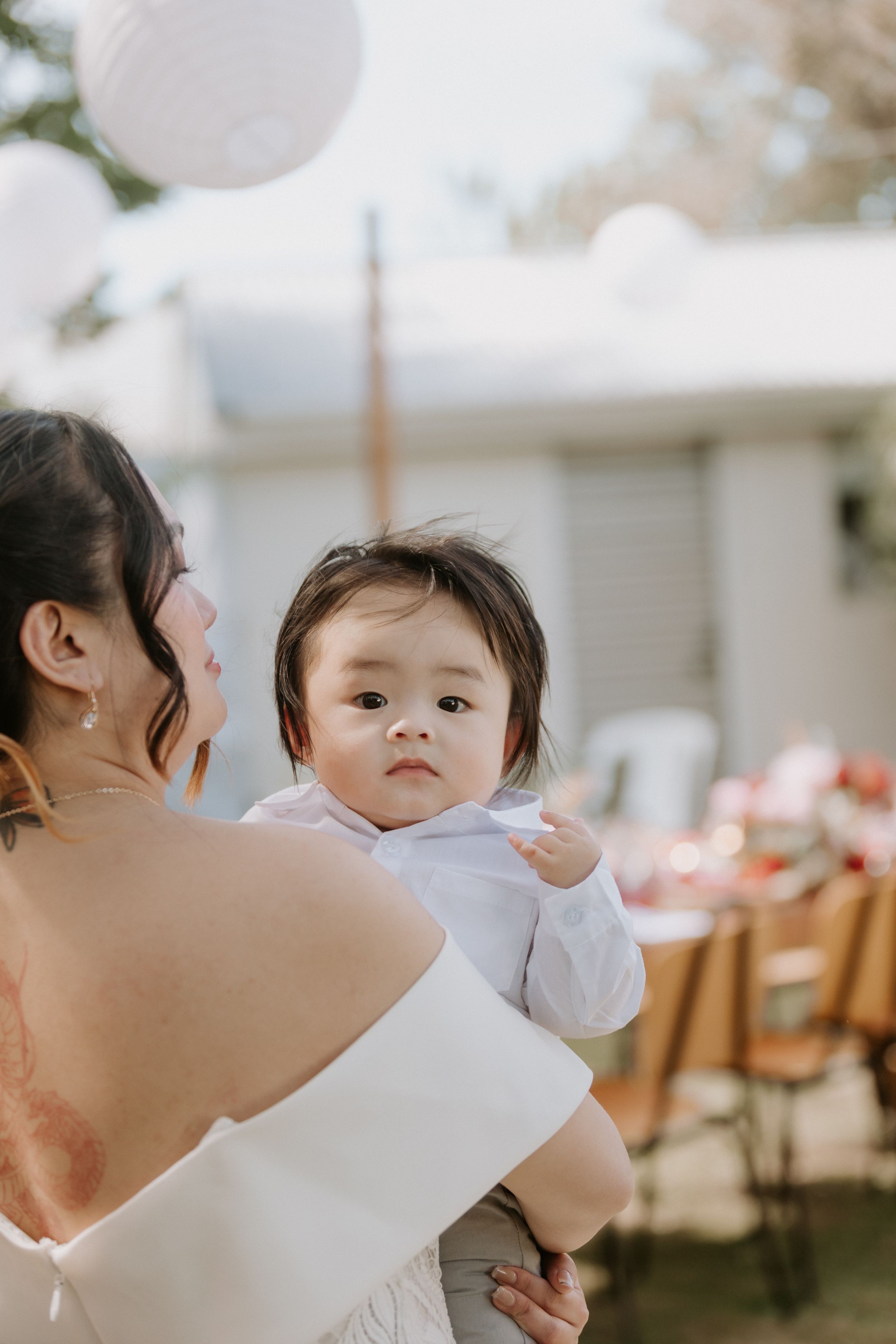 A parent holds and kisses a baby in white clothing at an outdoor event with tables in the background.