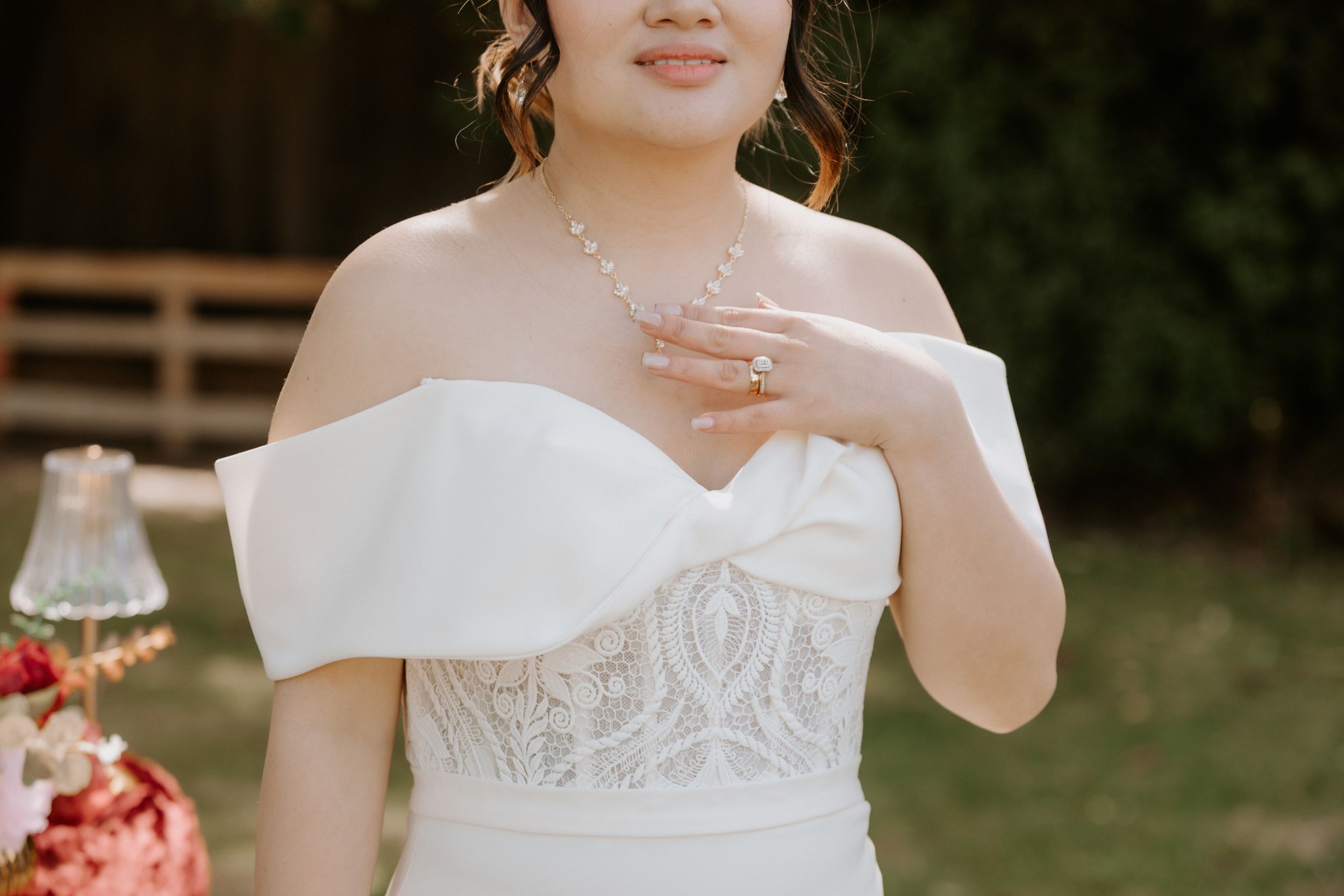 A bride in an elegant off-shoulder white wedding gown poses in a garden setting with delicate floral decor.