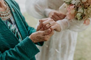 Close-up of hands exchanging wedding rings during ceremony with emerald green sweater and floral bouquet in background.