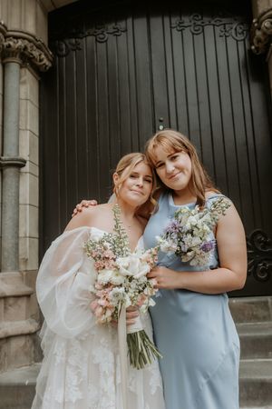 Two people in wedding attire embrace while holding a bouquet of pink and white flowers against a dark background.