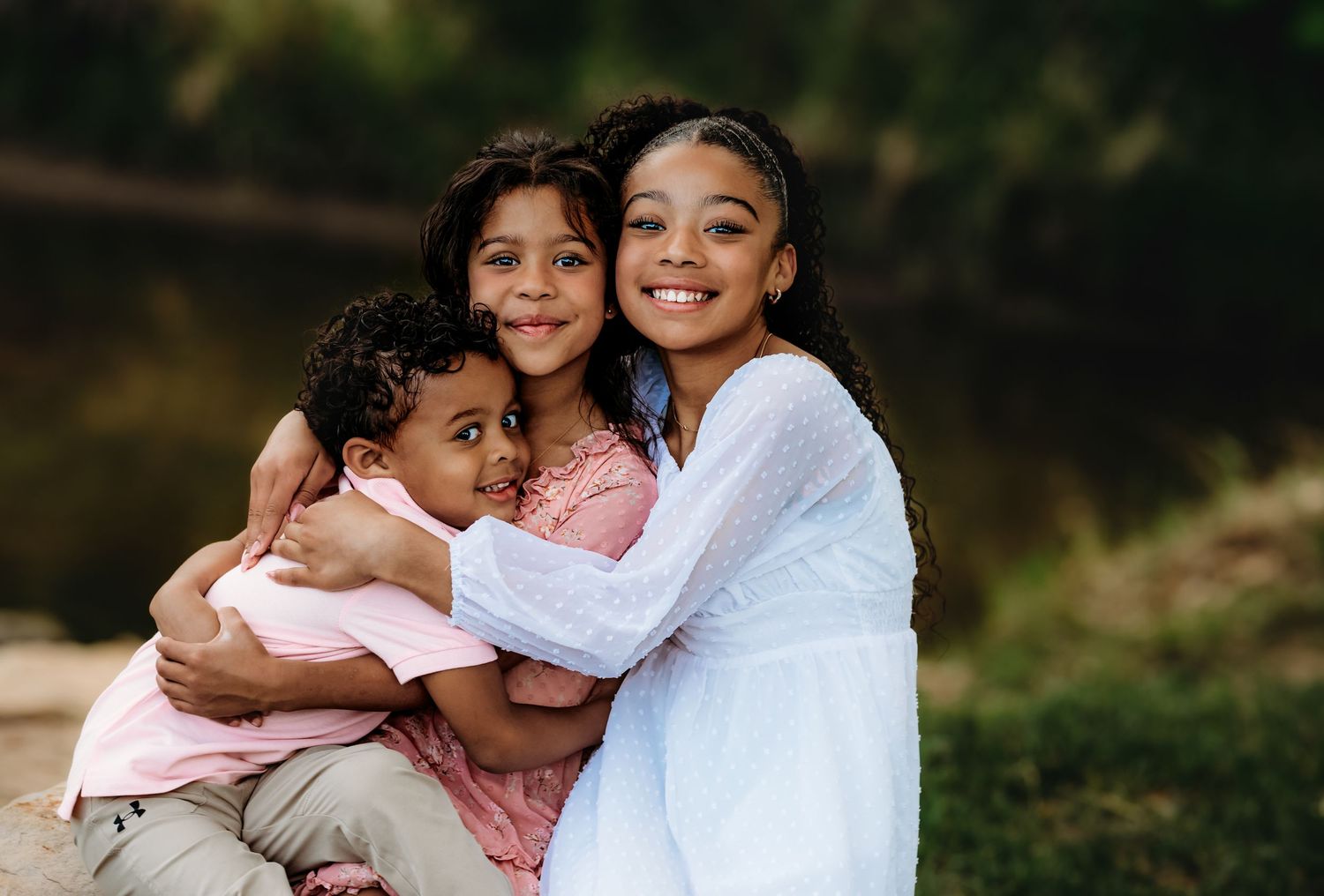 Family members share tender embraces while wearing white and pink clothing in an outdoor setting.