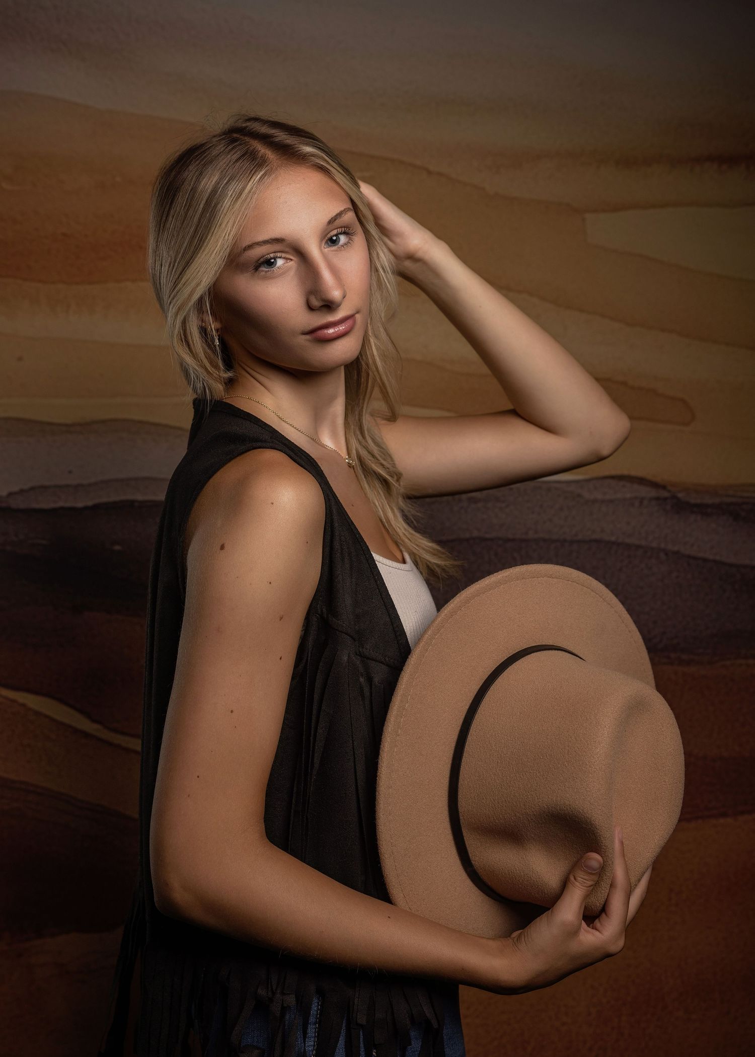 A person in jeans and boots sits on wooden steps holding a tan fedora hat in a moody studio portrait series.