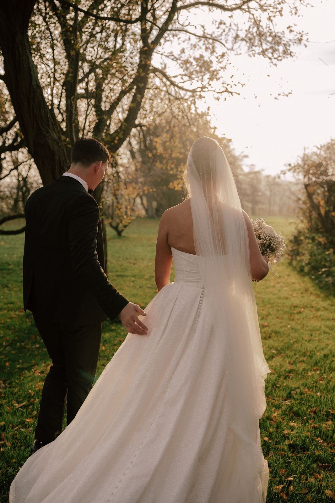 A couple walks through a sunlit autumn field with the bride's white wedding dress flowing in the gentle breeze.