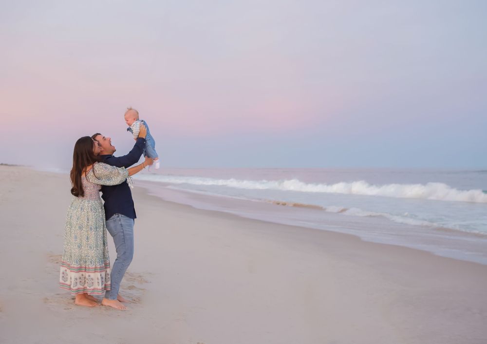 A person lifts a baby in the air while standing on a beach at sunset with ocean waves in the background.