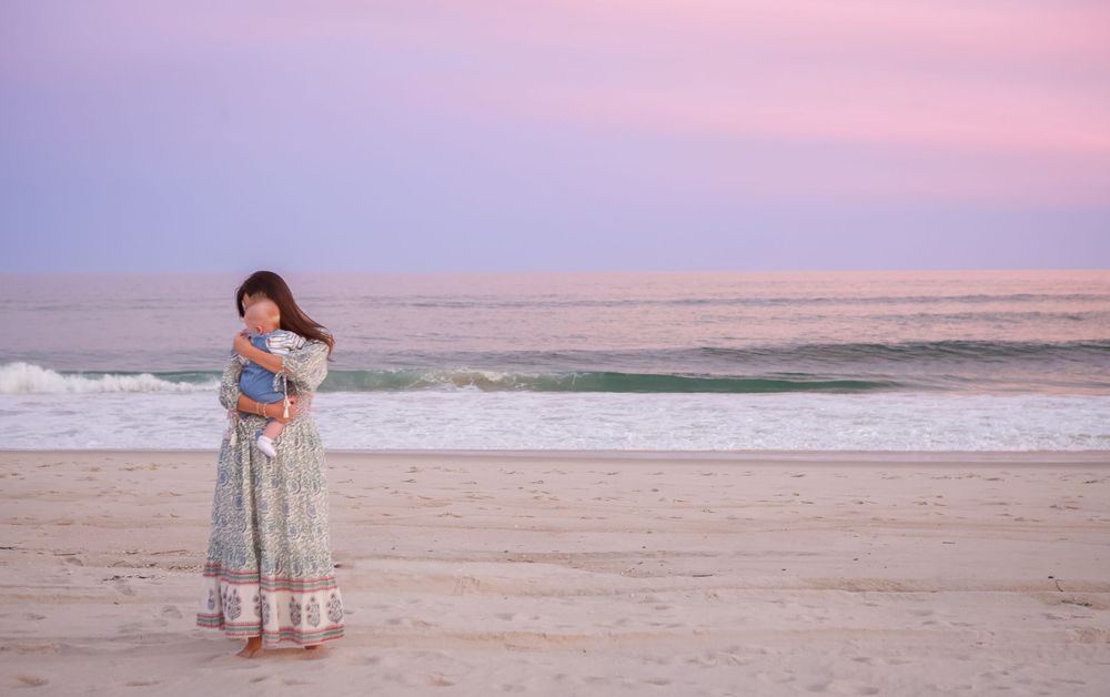 A person walks along a peaceful beach at sunset with soft pink skies reflecting on the sand and water.
