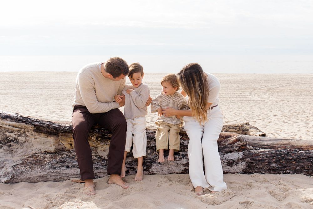 A family in neutral-colored clothing shares a tender moment together on a sandy beach at sunset.