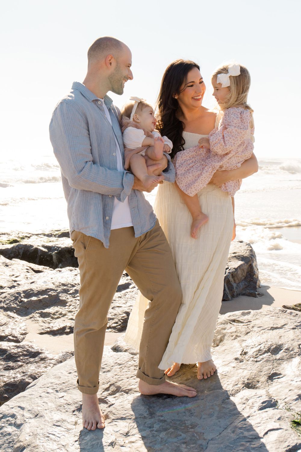 Family shares tender moments together on a sunny beach with rocky outcrops in the background.