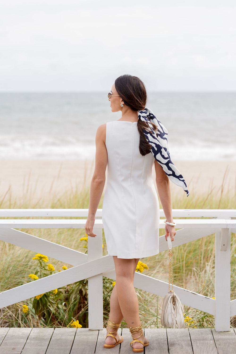 Child in white dress and sandals walks along coastal boardwalk with beach grass and ocean in background.
