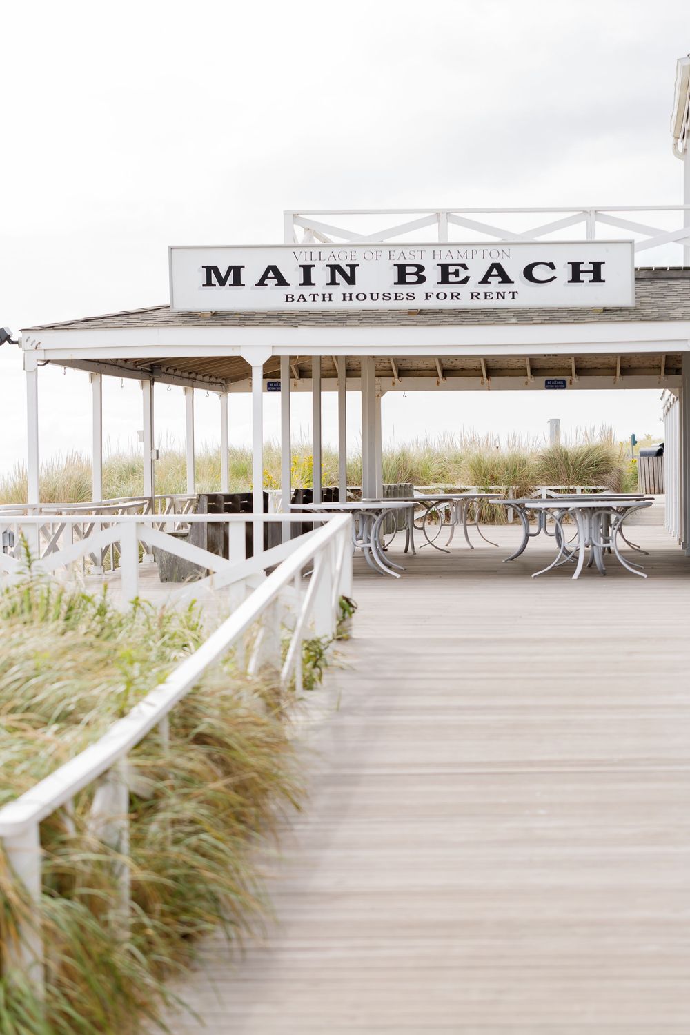 Wooden boardwalk leading to Main Beach entrance with coastal grass and white railings on a cloudy day.