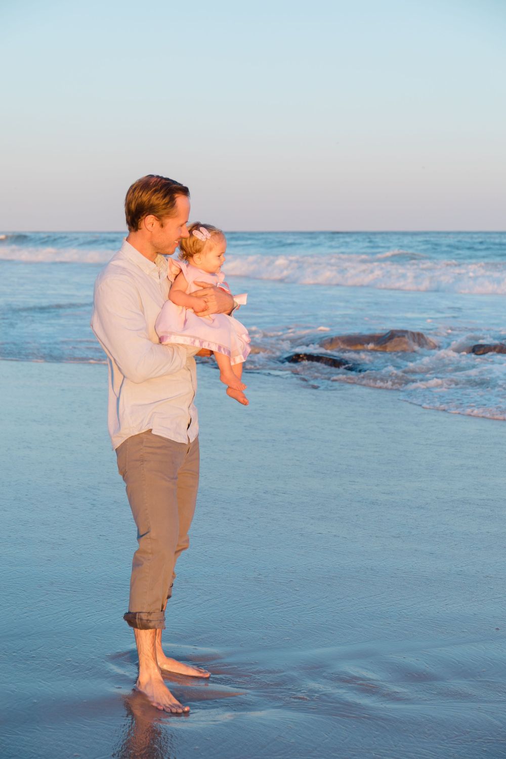 Parent and child playing in the shallow ocean waves at sunset with rocks visible in background.