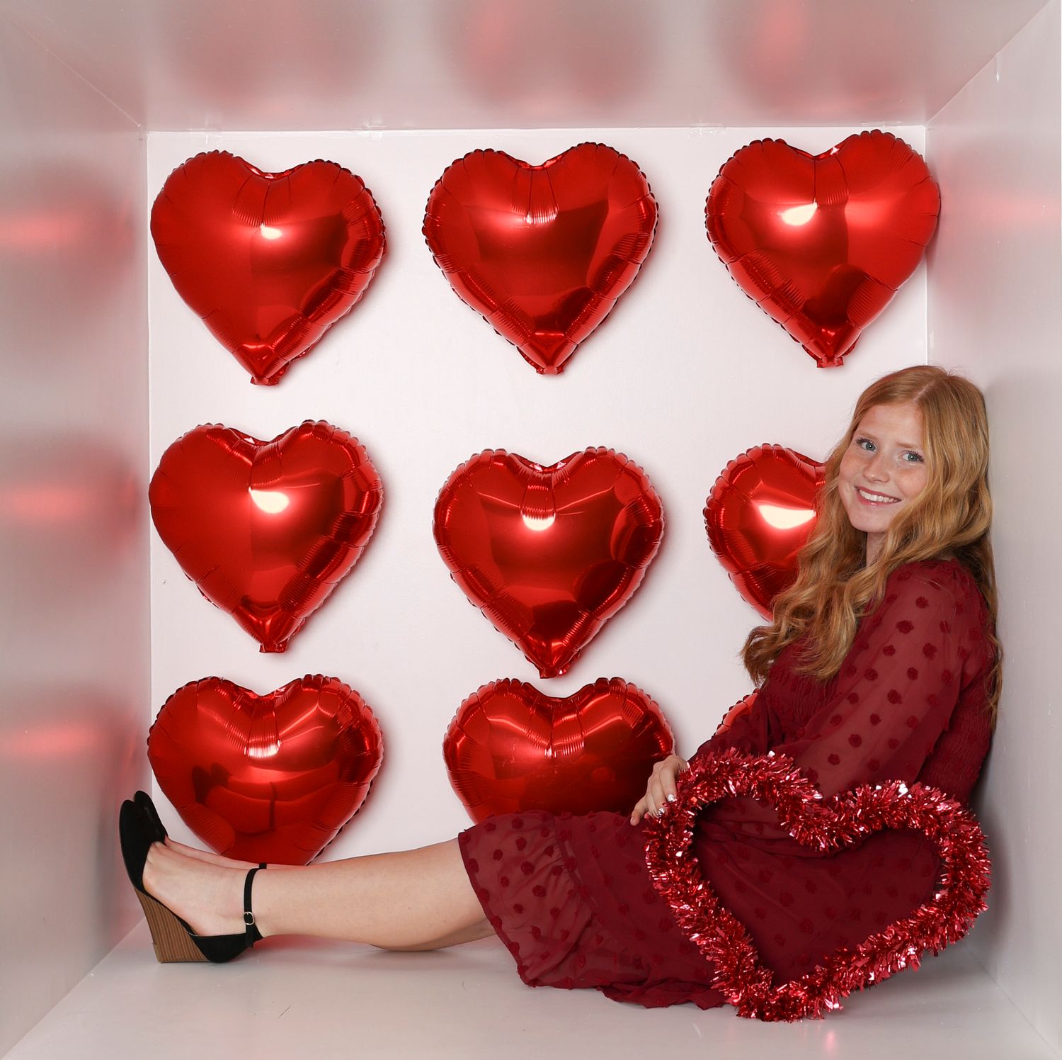 Valentine's themed photo shoot setup with red heart balloons and a person in a burgundy dress against a white backdrop. Box photography