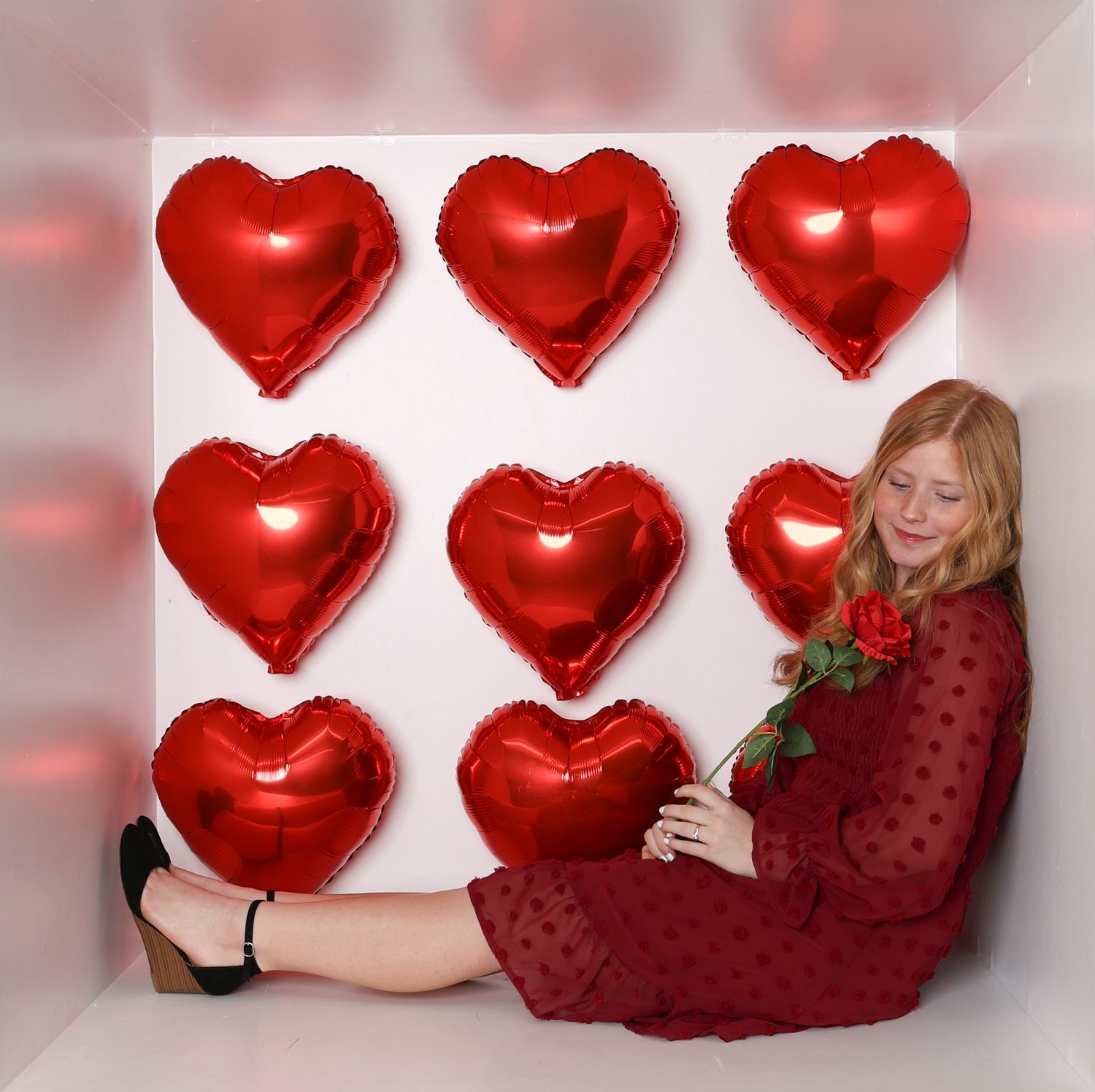 Romantic Valentine's scene with red heart balloons and someone wearing a burgundy dress holding a rose. Box photography session