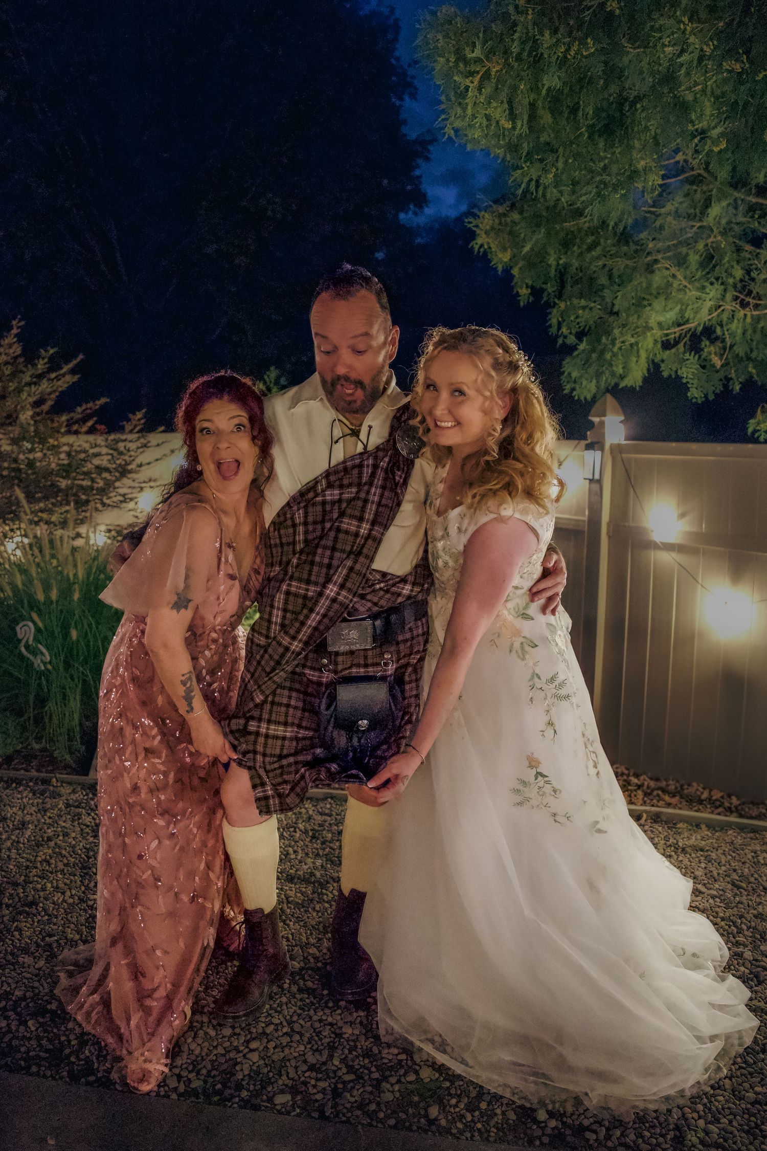 Three people share a joyful moment laughing together during an outdoor evening celebration in formal attire.