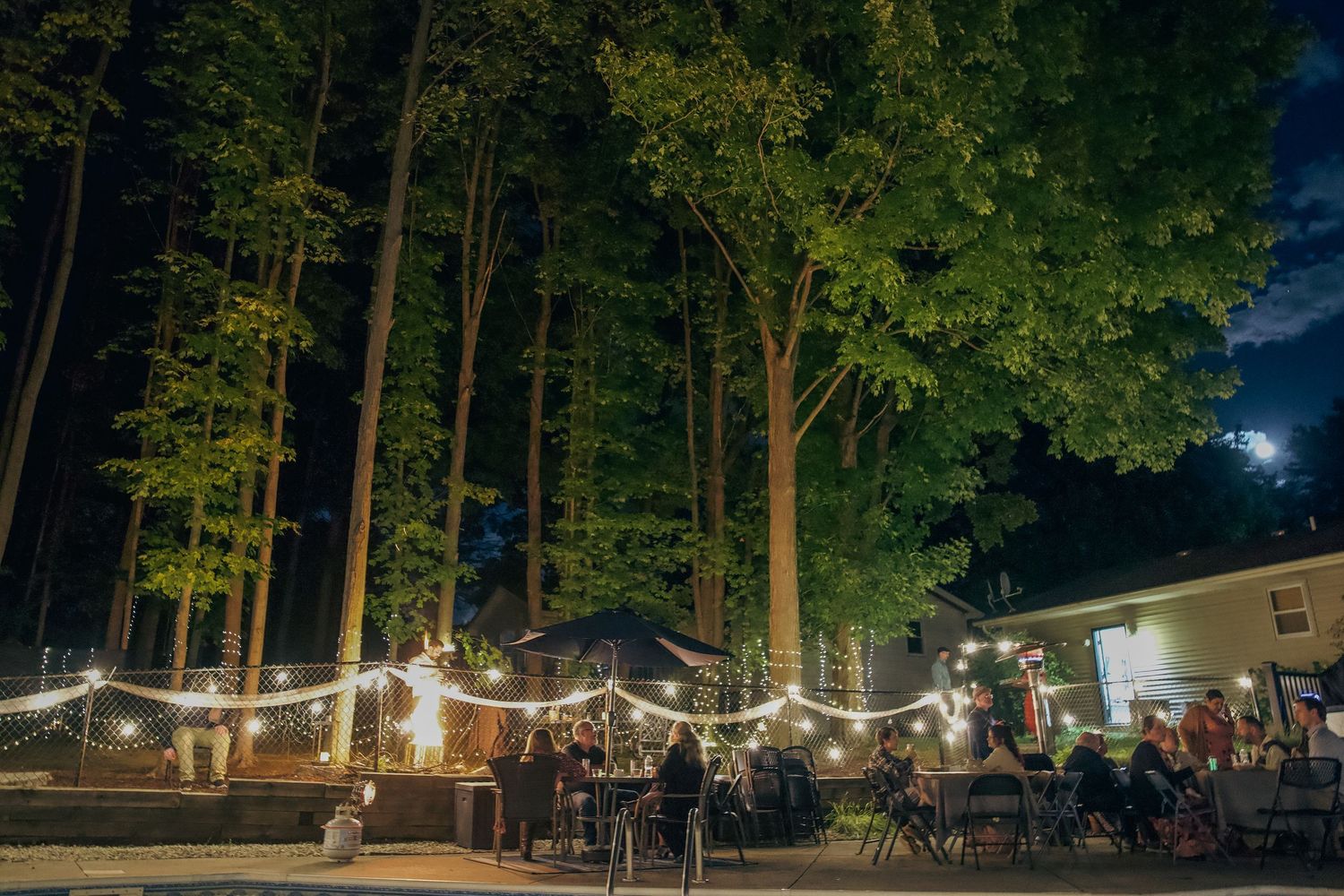 Outdoor event venue at night with tall pine trees illuminated by string lights and scattered tables and chairs.