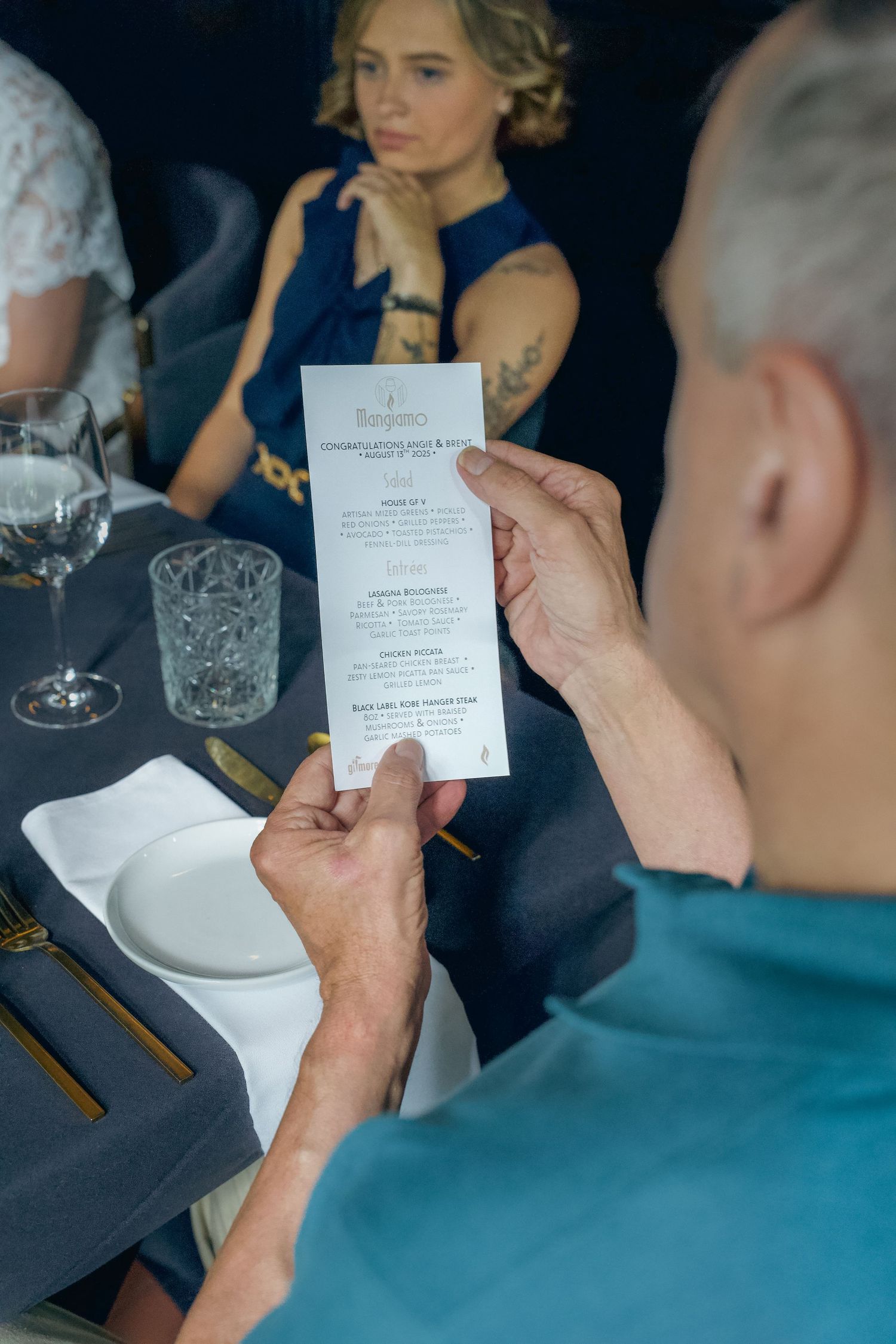 A person in a teal shirt reads a menu at an elegant dinner table with crystal glassware and place settings.