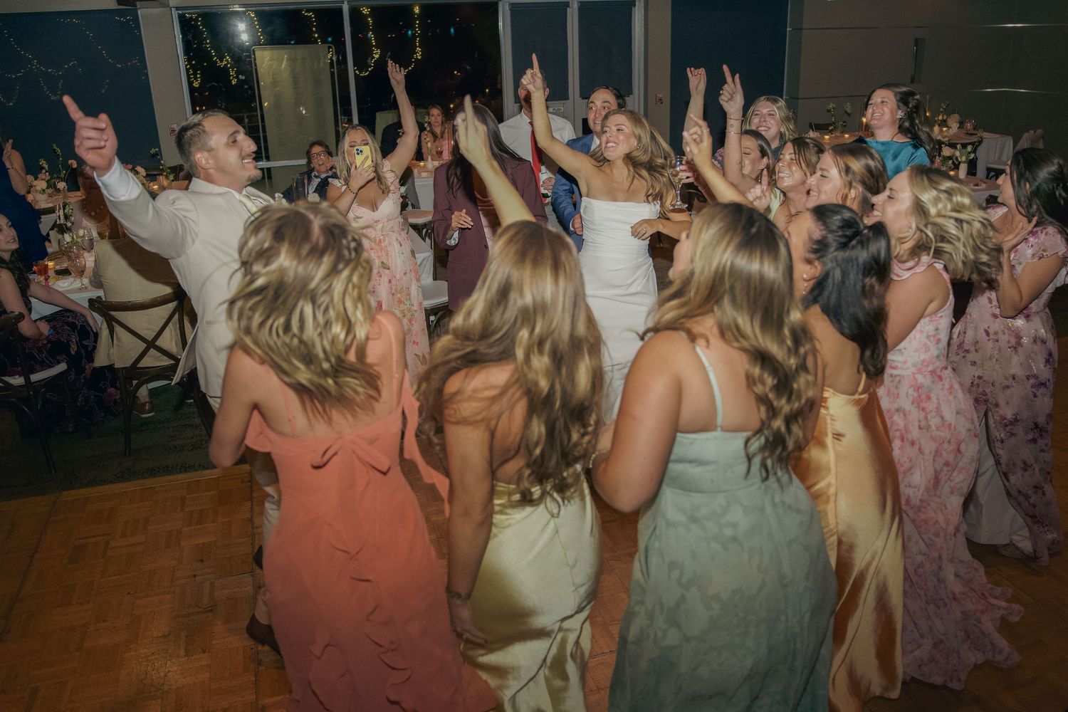Wedding guests dance and celebrate together on a dimly lit dance floor at an evening reception.