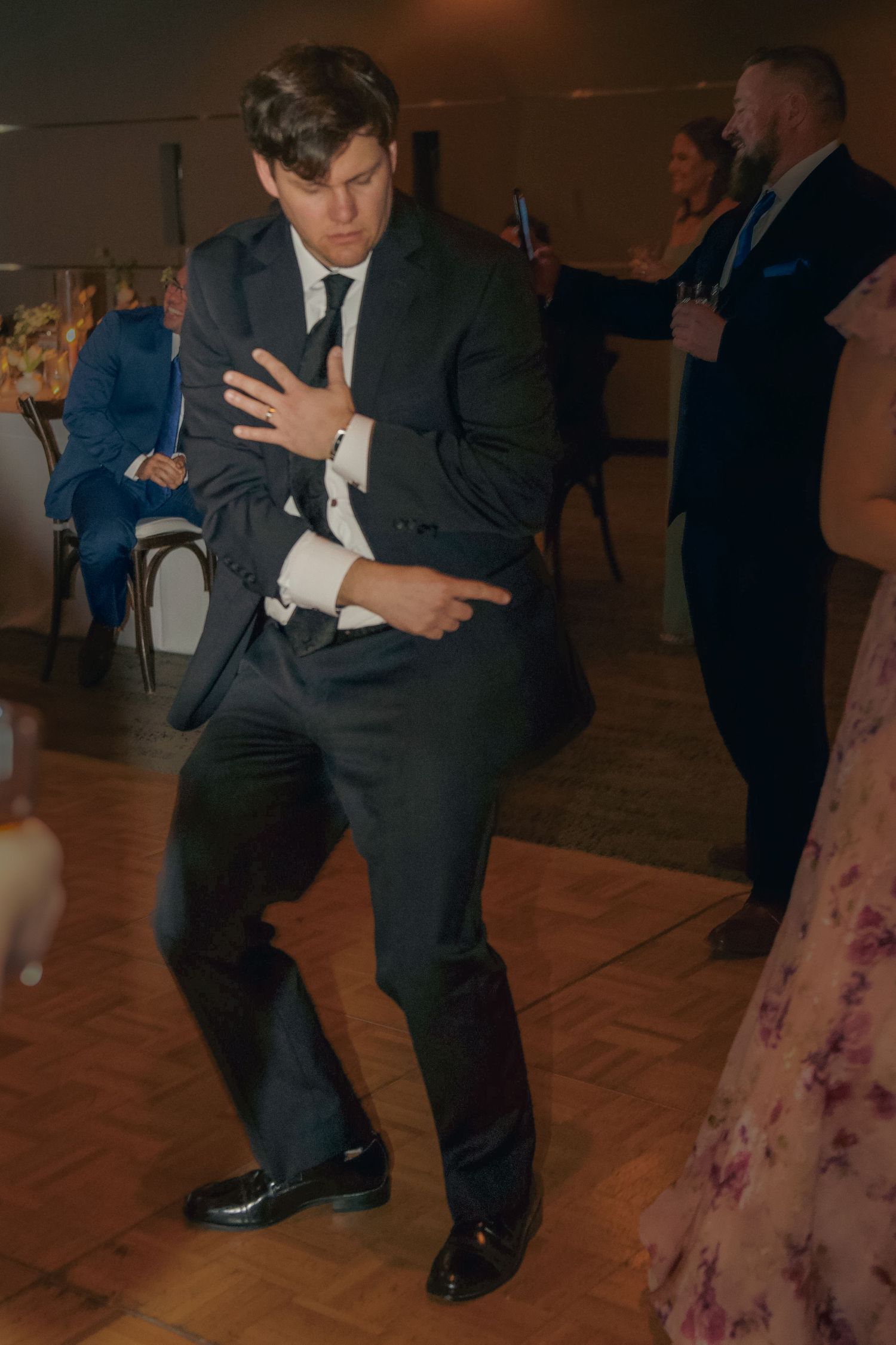 Person in a dark suit performs dance moves on a wooden floor at what appears to be a formal event.
