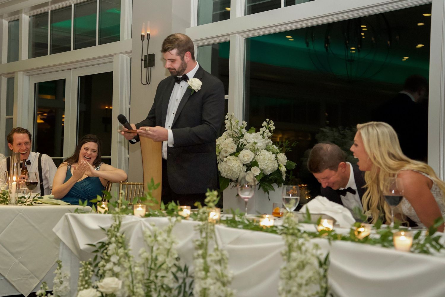 Wedding guests enjoy an elegant dinner reception at tables decorated with white floral centerpieces and chandeliers.