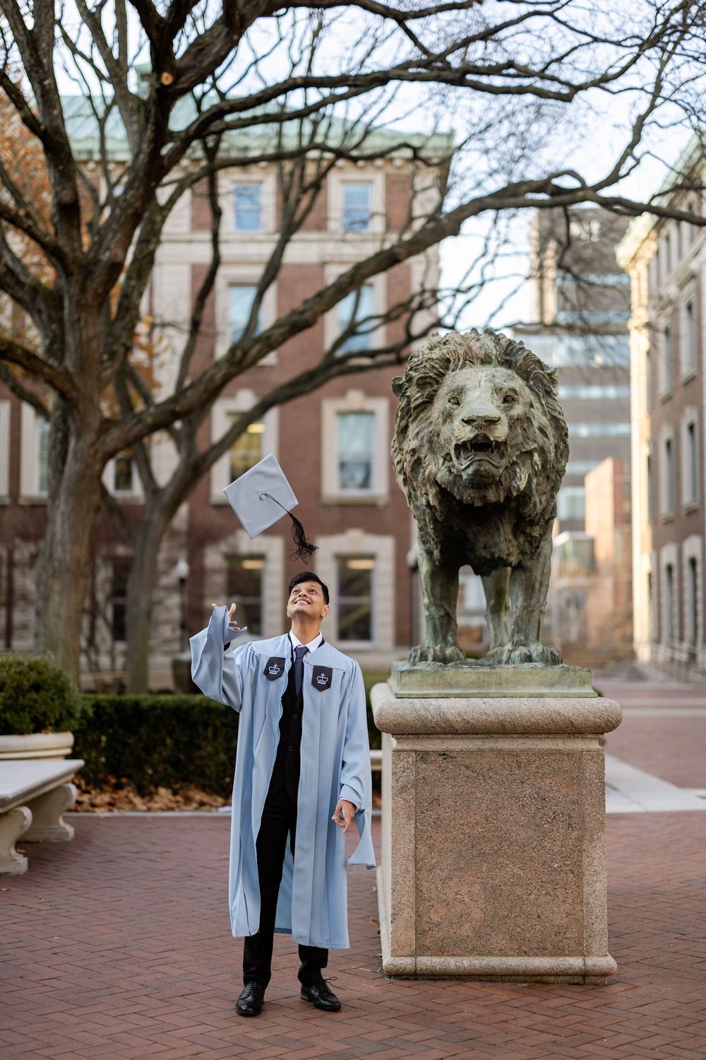 A graduate in light blue academic regalia celebrates on a brick walkway surrounded by historic university buildings.