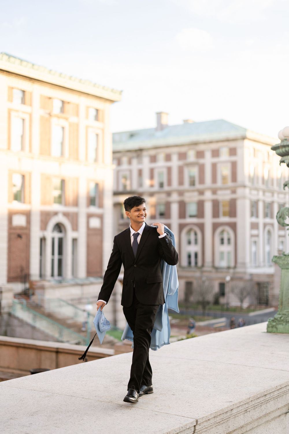 A graduate in a black suit walks with a diploma and umbrella on a university campus with historic buildings in the background.