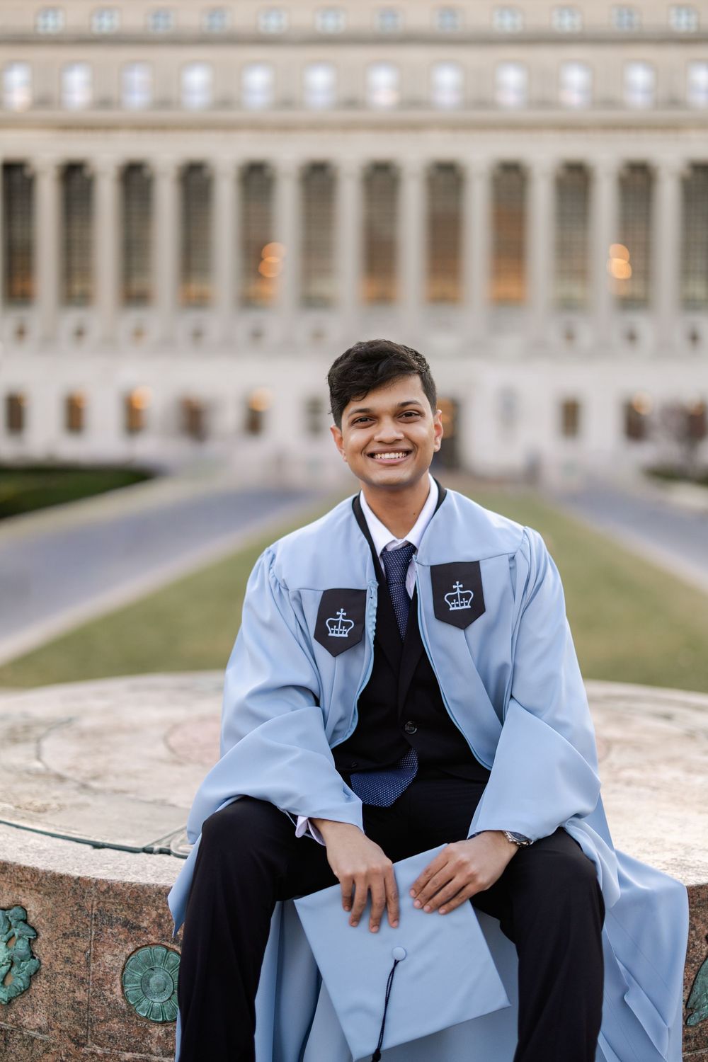 Graduate in light blue academic regalia sits on stone ledge in front of columned university building at dusk.