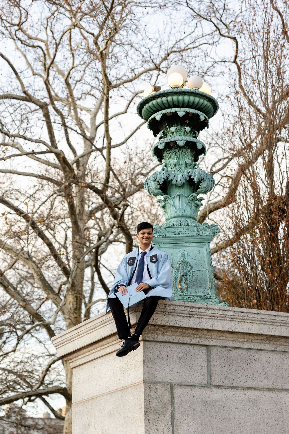 A student in academic regalia sits atop an ornate green bronze lamppost pedestal on a university campus.