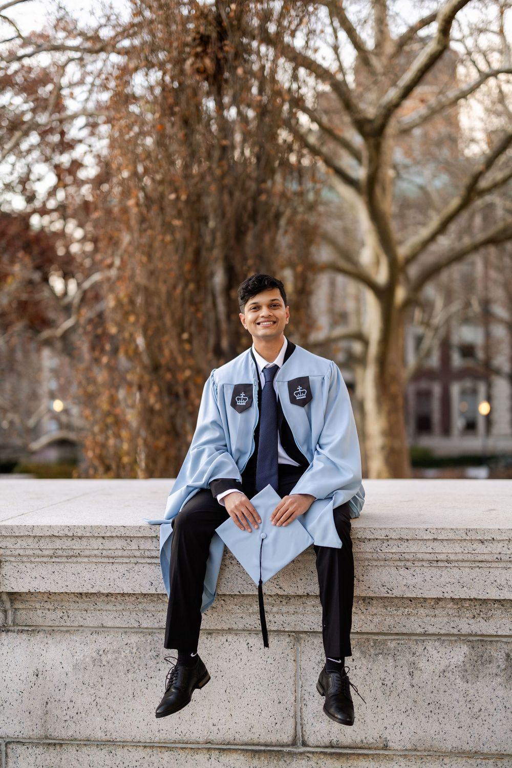 Graduate in light blue regalia sits casually on stone wall with autumn trees in background.