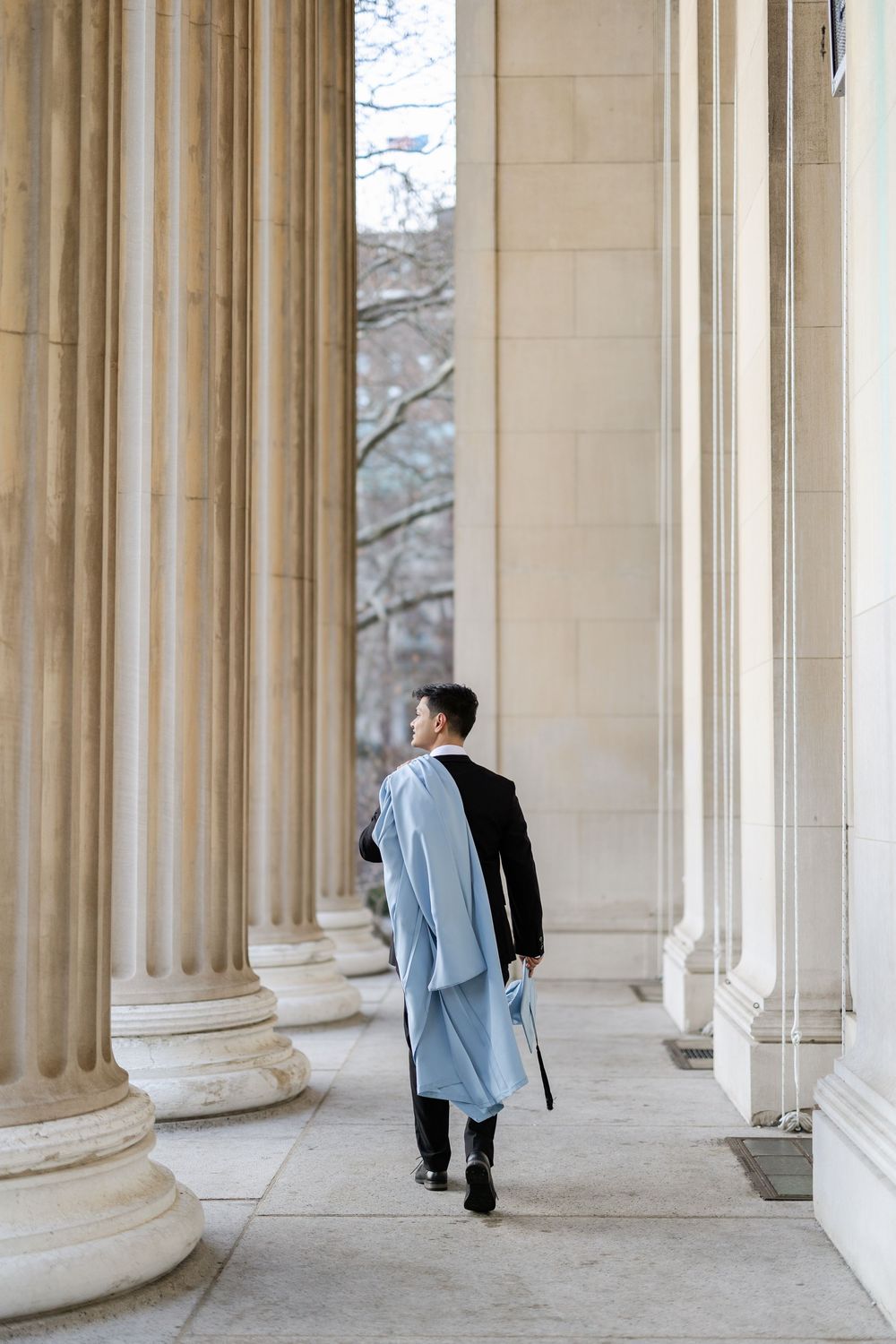 Graduate in light blue regalia walks through classical stone colonnade on university campus.