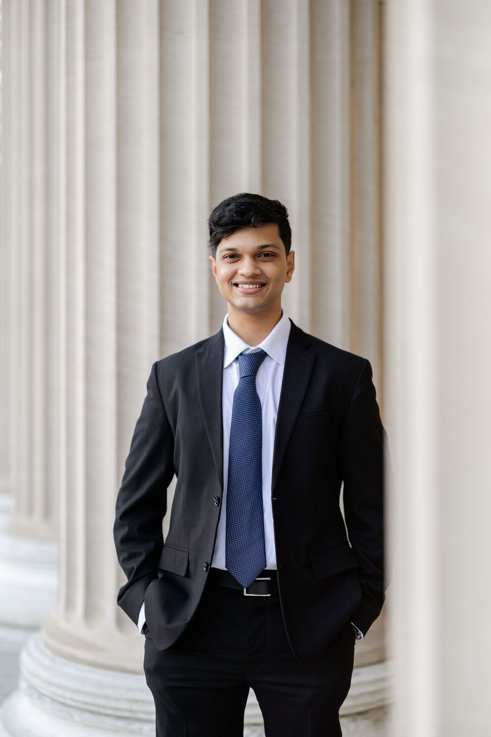 Professional portrait in dark suit and blue tie standing in front of white columns.