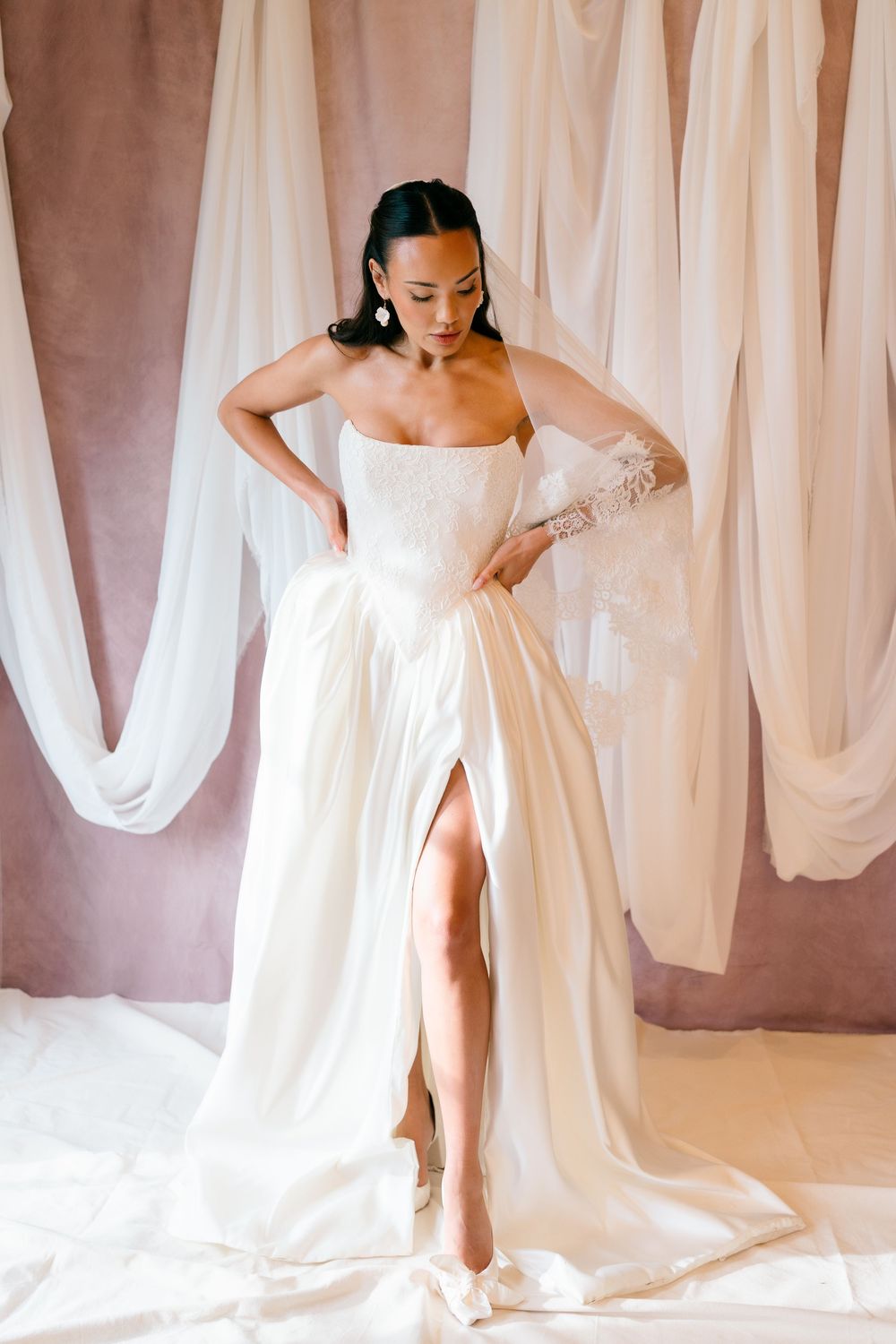 Elegant strapless white wedding gown with high slit and flowing train against draped fabric backdrop.