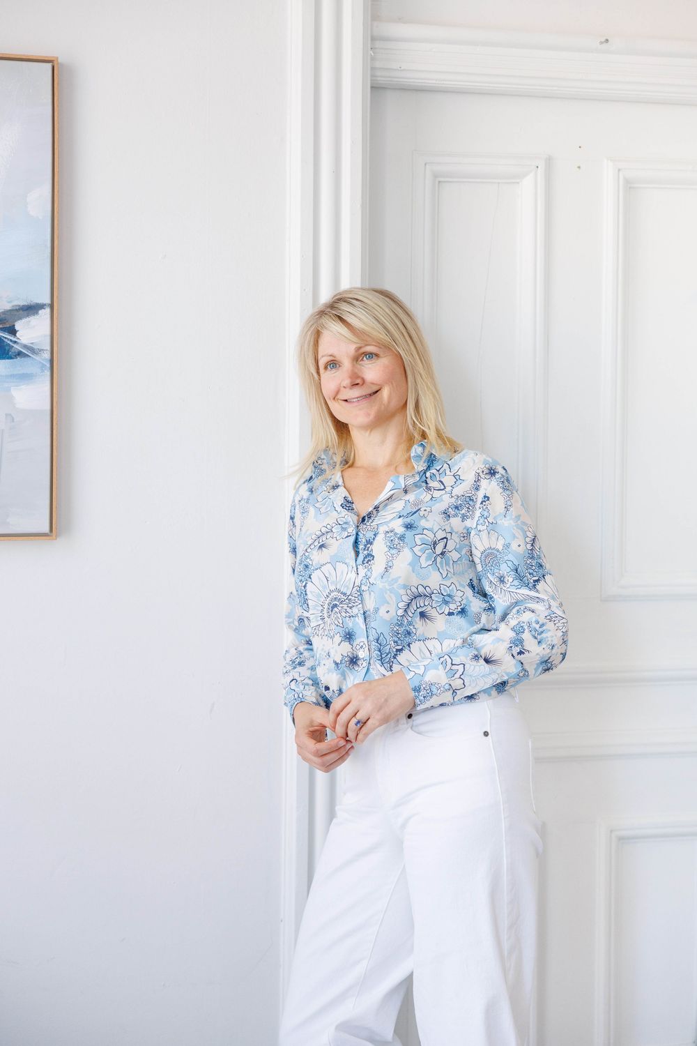 Person in blue and white floral top and white pants poses against white wall in bright interior setting.